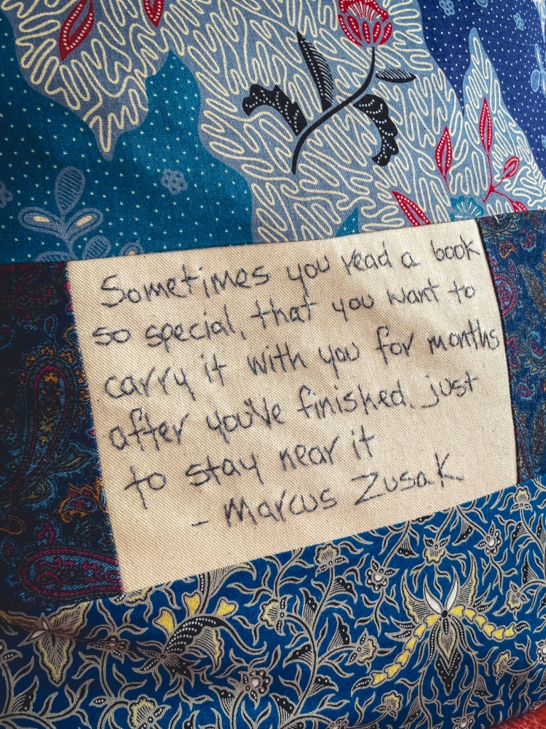 [Image Description: Blue fabric cushion with embroidery writing that says 'Sometimes you read a book so special, that you want to carry it with you for months after you've finished, just to stay near it - Marcus Zusak'.]