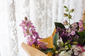 [Image Description: A bunch of mixed flowers, wrapped in brown paper with a white lace curtain in the background.]