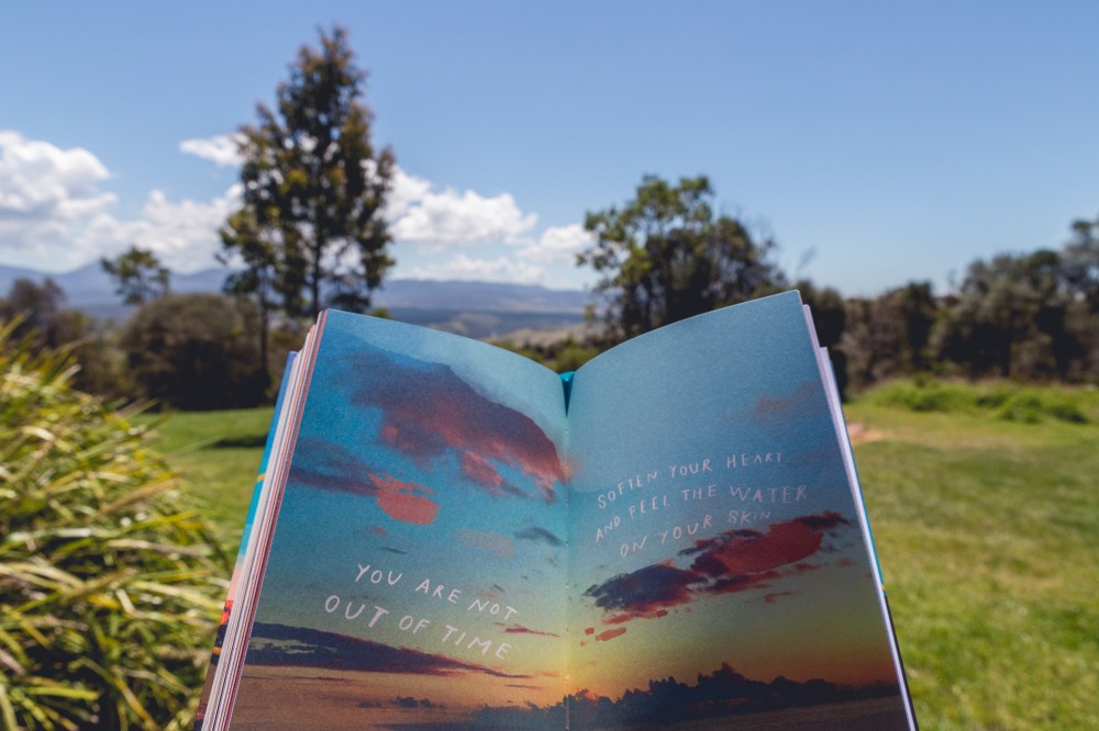 [Image Description: Morgan Harper Nichols' book open to page with sunset coloured sky and clouds and the words "You are not out of time. Soften your heart and feel the water on your skin". In the background is grass, shrubs and trees with blue mountains and sky in the far distance.]