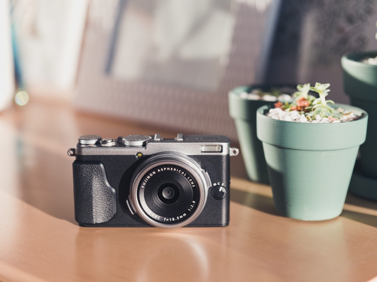 [Image Description: small retro style black and silver camera on a timber surface with small green pot plants and white picture frames in the background.]