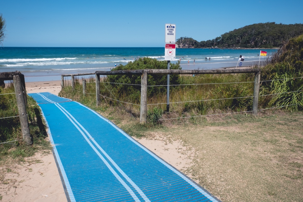 [Image Description: a bright blue mobility mat stretches out into the distance from the grass area to the beach. Timber railings, plants and signs in front of a bright Australian beach with sunshine, flags and swimmers.]